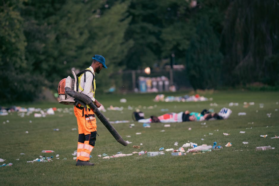 A sanitation worker dressed in a high-visibility orange trousers and a reflective safety vest, wearing a blue cap, is operating a leaf blower on a grassy park area. The worker is positioned on the left side of the image, with the blower's tube directed towards the ground, clearing scattered litter including plastic bottles, paper, and other debris. In the background, there is a person lying on the grass, and multiple trash items are visible strewn across the scene. The environment features lush green trees and shrubbery, with a few indistinct structures and a waste disposal area in the distance. The lighting suggests late afternoon or early evening, casting soft natural light over the area. The scene demonstrates an organized effort in waste collection and clean-up, aligning with professional rubbish removal services such as those provided by Rubbish Removal Belsize Park, for maintaining outdoor spaces free from litter and debris.