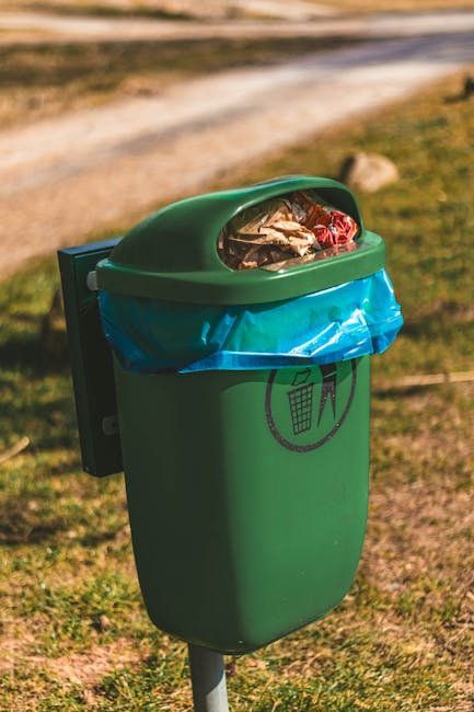A green outdoor rubbish bin with a rounded lid, positioned on a metal stand on a grassy area with patches of soil. The lid is slightly open, revealing assorted waste including crumpled paper, plastic wrappers, and debris inside, which are visible through a transparent section on the top. A blue plastic bag lines the interior of the bin, covering its sides and bottom. In the background, a dirt path stretches horizontally across the image, with blurred greenery and rocks beyond, indicating an outdoor environment. The scene is illuminated by natural daylight, highlighting the textures of the waste and the bin's smooth surface. The bin features a black recycling symbol printed on the front, and its presence suggests a focus on outdoor waste management, possibly related to private rubbish collection services like those offered by Rubbish Removal Belsize Park. The overall setting appears clean and organized, with a subtle connection to alternative waste handling outside traditional municipal collection methods.