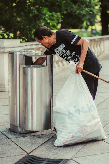 A middle-aged man with dark hair, dressed in a black t-shirt with white text and dark trousers, is seen standing on a paved sidewalk in an outdoor setting with lush green trees in the background. He is holding a white plastic bag filled with rubbish in his left hand and using a wooden stick or grabber with his right hand to place loose waste into a cylindrical silver metal litter bin that features two openings and is positioned next to a stone balustrade. The scene is well-lit by natural daylight, and the overall environment suggests a clean, well-maintained public space where independent or community-led rubbish collection activities might be taking place. The man appears to be engaged in waste disposal or rubbish removal, reflecting services offered by companies like Rubbish Removal Belsize Park for managed local cleaning and rubbish clearance projects that focus on private waste handling outside of standard municipal services.
