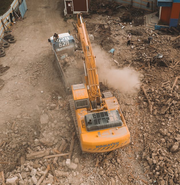 An aerial view of a construction site showing a large yellow excavator with the brand name 'HEAVY'S U' on its side, actively engaged in rubble removal. The excavator's arm extends forward, gripping and moving broken bricks, concrete debris, and other demolition materials. Dust and small particles are visible as the machine works on the uneven, debris-strewn ground, which is covered with scattered concrete blocks, wooden planks, and loose soil. To the left, a section of the site is enclosed with a temporary fence, and beyond that, a few construction workers are visible standing among the rubble. In the background, part of a partially demolished structure with exposed walls and scattered fittings can be seen, indicating ongoing demolition or site clearance activities. The scene is captured in natural daylight, highlighting the textures of the dusty environment and the rugged construction debris, with a focus on the machinery that supports efficient waste removal and site clearing, aligning with private rubbish collection and on-site clearance services offered by Rubbish Removal Belsize Park.