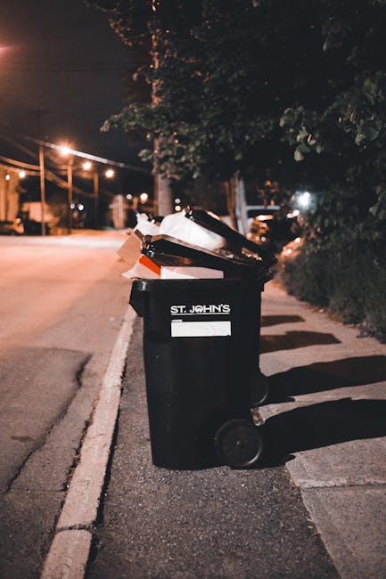 A black wheeled rubbish bin with a white label and the text 'ST. JOHN'S' printed on it is positioned on the sidewalk near the curb, filled with a variety of waste materials including cardboard boxes, plastic containers, and paper bags. The bin is placed on a paved pavement with a white line marking the edge of the road. In the background, a tree casts shadows over the scene, illuminated by warm streetlights that create a soft glow against the dark night sky. Additional bins are visible further down the sidewalk, suggesting regular waste collection or a private waste disposal service. The environment appears residential, with parked cars, utility poles, and a quiet street extending into the distance. The scene reflects an ordinary nighttime setting for rubbish collection, potentially managed by an independent waste removal service like Rubbish Removal Belsize Park, supporting alternative waste handling options outside of council services.