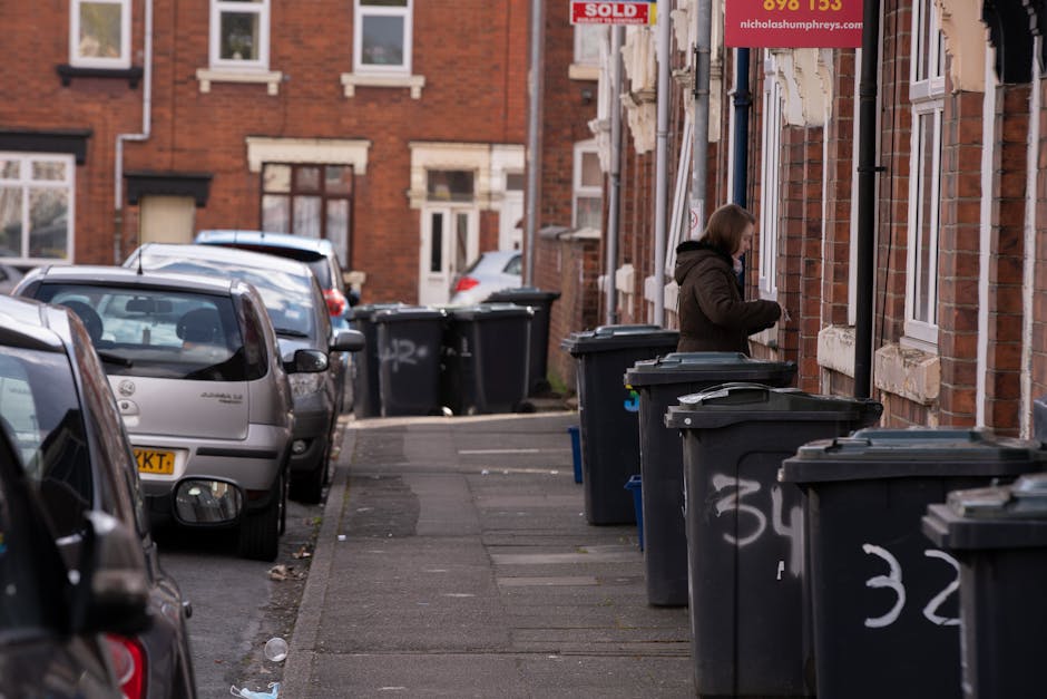 The image shows a residential street lined with traditional red-brick terraced houses, each with white-framed windows and stone detailing around the openings. On the left side of the image, several parked cars, including a silver compact car closest to the foreground, occupy the street along the curb. The pavement features a narrow sidewalk with a slightly uneven surface, scattered with small debris, and is lined with multiple black wheelie bins with closed lids, some labelled with white spray paint numbers, positioned along the front of the houses. A woman, dressed in a dark hoodie and jacket, stands near one of the bins, appearing to be in the process of discarding rubbish or organising waste. The background reveals more parked vehicles and additional residential buildings, with some signage indicating real estate activity. The overall environment is illuminated by natural light, possibly during daytime, with no visible ongoing activity but a clear emphasis on waste disposal, highlighting the local practice of private rubbish collection in the area, as often managed by rubbish removal services such as Rubbish Removal Belsize Park.